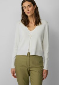 Witte cardigan met geribde manchetten en zoom, v-hals, sluiting met één knoop, gedragen over een lichte top en olijfgroene broek.