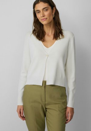 Witte cardigan met geribde manchetten en zoom, v-hals, sluiting met één knoop, gedragen over een lichte top en olijfgroene broek.