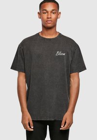 Camiseta negra de gran tamaño con acabado de denim desgastado, que presenta el texto bordado "Bloom" en el lado izquierdo del pecho. Mangas cortas y cuello redondo.