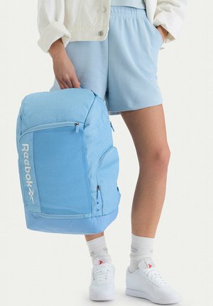 Osoba trzymająca jasnoniebieski plecak Reebok, ubrana w jasnoniebieskie szorty, białe skarpetki, białe tenisówki i białą kurtkę na białym tle.