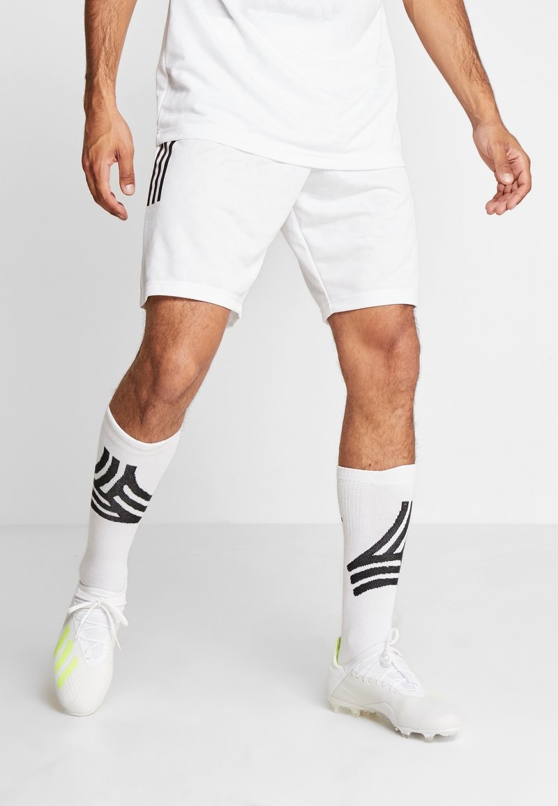 Athlète masculin portant un short de sport blanc, des chaussettes blanches hautes avec des rayures noires, et des chaussures à crampons blanches avec des détails vert néon.