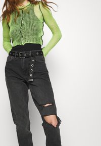 Top de malla verde de manga larga con cuello redondo, combinado con vaqueros negros de tiro alto desgastados que presentan una cintura con cinturón y hardware metálico.