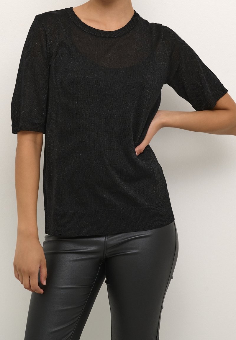 Femme portant un haut en tricot noir à manches courtes avec un décolleté transparent et un pantalon en simili cuir noir, posant avec une main sur la hanche.