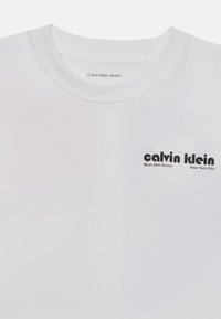 Bílá bavlněná tričko s kulatým výstřihem a krátkými rukávy. Černý textový logo s nápisem "calvin klein" a podrobnostmi o místě je umístěno na přední straně.