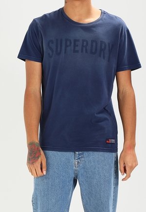 Personne portant un t-shirt Superdry bleu marine et un jean bleu clair avec un tatouage d'une rose rouge sur la main gauche.
