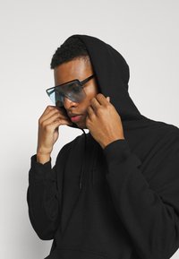 Sort hætte sweatshirt med en struktureret overflade. Personen har store, klare solbriller med vinklede stel og justerer hætte-snore.