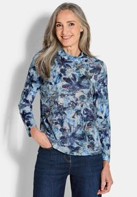 Langärmliges Oberteil in Hellblau mit floralem Muster in dunkleren Blautönen und goldenen Akzenten. Weiches Material, Rundhalsausschnitt, kombiniert mit dunklen Jeans.
