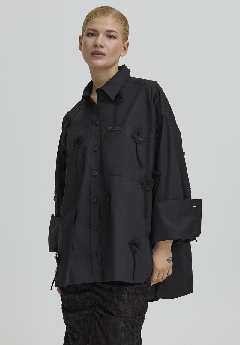 Kvinde iført løs sort skjorte med stofblomster og brede manchetter, kombineret med teksturerede sorte bukser, stående mod en ensfarvet baggrund.