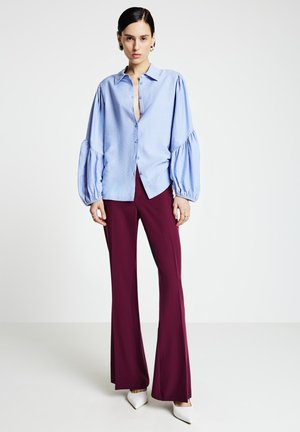 Woman wearing a light blue balloon-sleeve blouse, burgundy flared pants, and white pointed-toe heels standing on a plain white background.