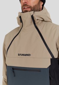 Bežová puloverová bunda s vysokým límcem, černými akcenty, velkou přední kapsou a odolným materiálem; logo značky vyobrazeno na hrudi.