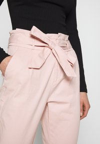 Ljust rosa byxor med bred midja och en rosettdetalj. Tillverkade av ett mjukt tyg, med sidofickor och en avslappnad passform.