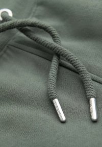 Tissu vert avec une texture lisse, présentant des cordons tressés se terminant par des embouts métalliques. Détail de couture subtil visible sur le tissu.