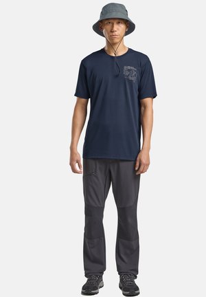 Homme portant un chapeau seau gris, un t-shirt graphique bleu marine, un pantalon gris foncé avec des genouillères, et des chaussures de randonnée noires, debout devant un fond blanc.