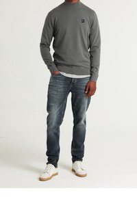 Grauer Langarm-Pullover mit Rundhalsausschnitt und Logo-Detail, kombiniert mit blauen Denim-Jeans und weißen Sneakers mit strukturierter Sohle.