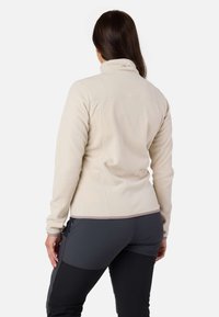 Beige fleece-takki, jossa on korkea kaulus, pitkät hihat ja ompeluyksityiskohtia. Yhdistetty tummanharmaisiin housuihin.