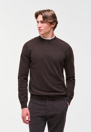 Giovane uomo con un maglione marrone scuro e pantaloni scuri, in piedi con una mano in tasca, che guarda verso sinistra su uno sfondo semplice.