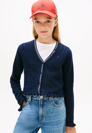 Tüdruk punase nokamütsi, tumesinise valgete äärtega nupuga kardigani, valge t-särgi ja kõrge vöökohaga siniste teksadega, seisab ühe käega taskus.