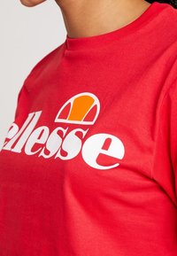 Camiseta de algodón roja con un gran logo blanco y un acento naranja y amarillo. Tiene mangas cortas y un diseño de cuello redondo. Textura suave.