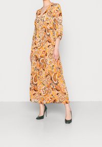 Kobieta w pomarańczowej sukience midi w wzór paisley z rękawami 3/4 i zielonych szpilkach, stojąca na jednolitym tle.