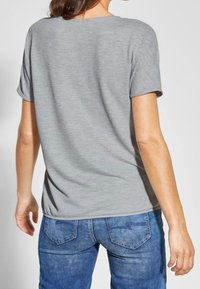 Szary top z krótkim rękawem, okrągłym dekoltem, teksturowanej tkaniny i luźnym kroju. Połączony z niebieskimi jeansami z tylnymi kieszeniami.