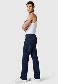Uomo con tatuaggi che indossa una maglietta bianca senza maniche, jeans blu scuro e sneakers bianche, in piedi con le braccia incrociate davanti a uno sfondo semplice.