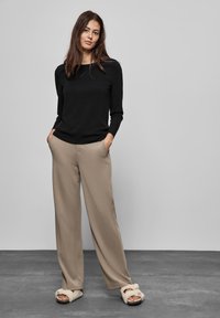 Zwarte blouse met lange mouwen en een ronde hals, beige wijde broek en fuzzy sandalen met open tenen. Model staat met de handen in de zakken.