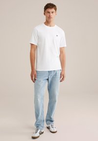 Mlad moški stoji obrnjen naprej, oblečen v navadno belo majico, svetlo modre jeans hlače in bele superge z črnimi stranicami.