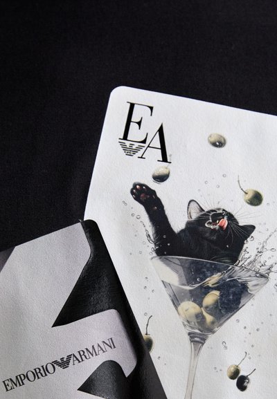 Fekete macska, amely egy martini pohárba nyúl, benne zöld olívabogyók, egy fehér lapon az "EA" logóval és az "EMPORIO ARMANI" szöveggel, fekete háttér előtt.