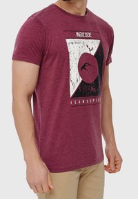 T-shirt in cotone borgogna con un design grafico che presenta una forma triangolare bianca, un elemento circolare e una texture invecchiata. Maniche corte.