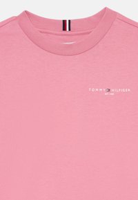 Roze kokvilnas T-krekls ar apaļu kaklu, uz kura ir balta "TOMMY HILFIGER" logo un 1985. gada izveides gads. Mīksta tekstūra, vienkāršs dizains.
