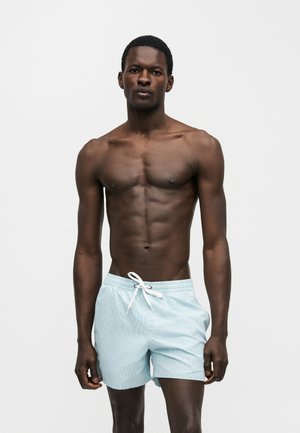 Muskuløs mand med mørk hud, iført lyseblå badebukser med snøre, stående mod en ensfarvet grå baggrund.