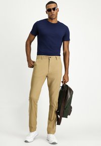 T-shirt a girocollo blu navy abbinato a pantaloni chinos beige. Il modello indossa occhiali da sole neri e tiene uno zaino verde con dettagli marroni. Le sneakers bianche completano il look.