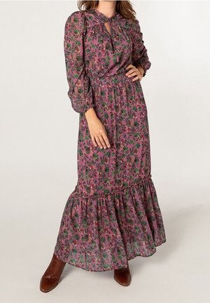 Mujer con un vestido largo morado con flores, mangas largas y botas marrones hasta el tobillo, de pie con una mano en la cintura.