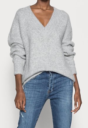 Personne portant un pull à col en V gris clair en laine duveteuse avec les manches retroussées et un jean bleu taille haute légèrement usé.