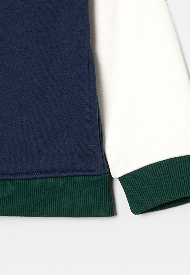 Sweatshirt bleu marine et crème avec des poignets côtelés verts. Tissu épais avec une texture lisse et un design de panneaux contrastants.
