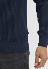 Temno modra teksturirana pulover s vzorcem vafljev, z rebrastimi manšetami in robom, ki ima na strani majhno oznako blagovne znamke.