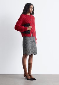 Roter gestrickter Cardigan mit Knopfverschluss; grauer Spitzenrock; braune gerippte Socken; schwarze Pumps mit Knöchelriemen; glatte, matte schwarze Clutch.