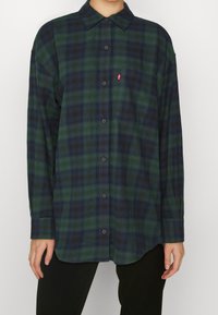 Grün und marineblau karierte Flanelljacke mit Button-Down-Kragen, langen Ärmeln und einer einzelnen Brusttasche, die ein kleines rotes Logo enthält.