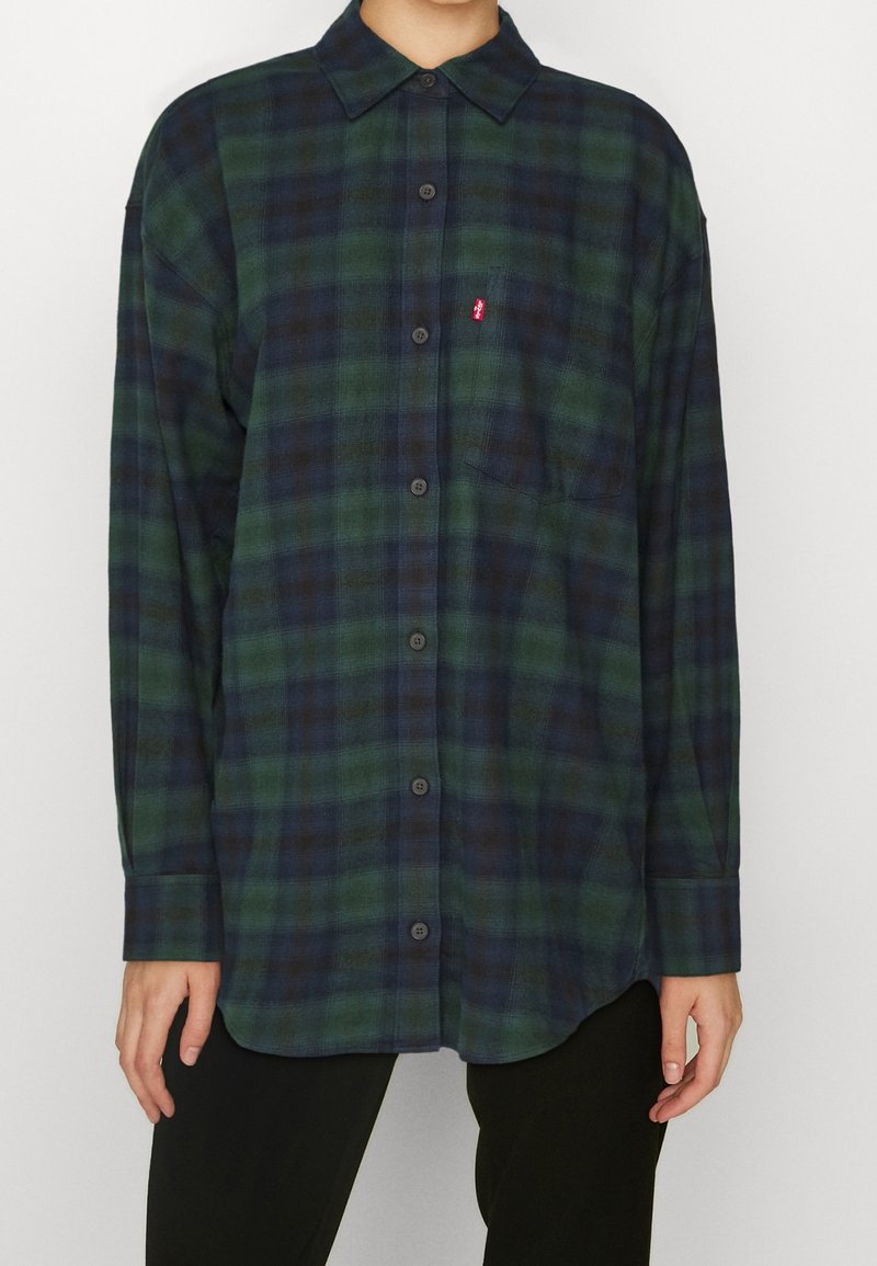 Grün und marineblau karierte Flanelljacke mit Button-Down-Kragen, langen Ärmeln und einer einzelnen Brusttasche, die ein kleines rotes Logo enthält.