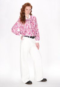 Lichte, doorzichtige, roze bloemen blouse met ritssluiting en witte manchetjes, gecombineerd met hoge, witte broeken en zwarte instapschoenen.