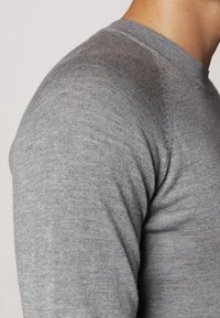 Grau farbener Rundhals-Pullover aus feinem Strickstoff, der eine glatte Textur und eine taillierte Passform zeigt. Stichdetails entlang der Schulternaht sind sichtbar.