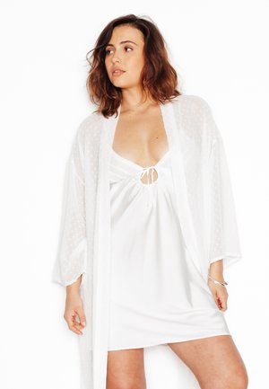 Femme aux cheveux bruns mi-longs portant une nuisette en satin blanc et un peignoir blanc transparent, regardant vers sa gauche.