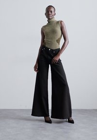Top verde cu nervuri, fără mâneci și cu un guler înalt, asociat cu pantaloni negri cu picioare largi. Modelul poartă tocuri negre. Fundal simplu.