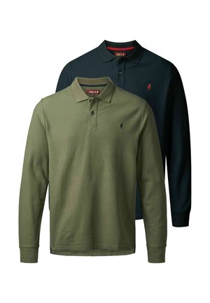 Homme avec barbe portant un polo à manches longues bleu marine foncé, un pantalon bleu marine et des bottes en cuir marron, debout devant un fond clair uni.