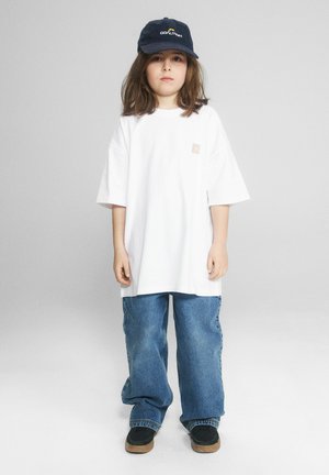 Dziecko stoi, nosząc czarną czapkę, oversize białą koszulkę, luźne niebieskie dżinsy i czarne buty na jednolitym szarym tle.