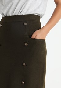 Mörkgrön stickad kjol med sidofickor, en åtsittande midja och dekorativa knappar fram för stängning. Slät textur.