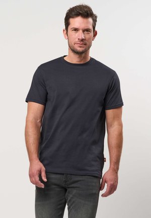 Muž s krátkymi hnedými vlasmi, oblečený v jednoduché čierne tričko s krátkym rukávom a tmavé džínsy, stojaci pred svetlým pozadím.