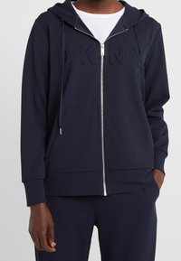 Marinblå zip-up hoodie i mjuk tyg, med en känguruficka, dragsko-huva och präglad "DKNY"-logga över bröstet.
