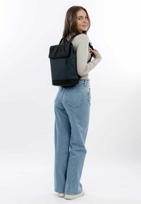 Sac à dos en tissu noir et marine avec un design à rabat, des bretelles ajustables et une finition texturée, porté sur le dos contre un fond clair.