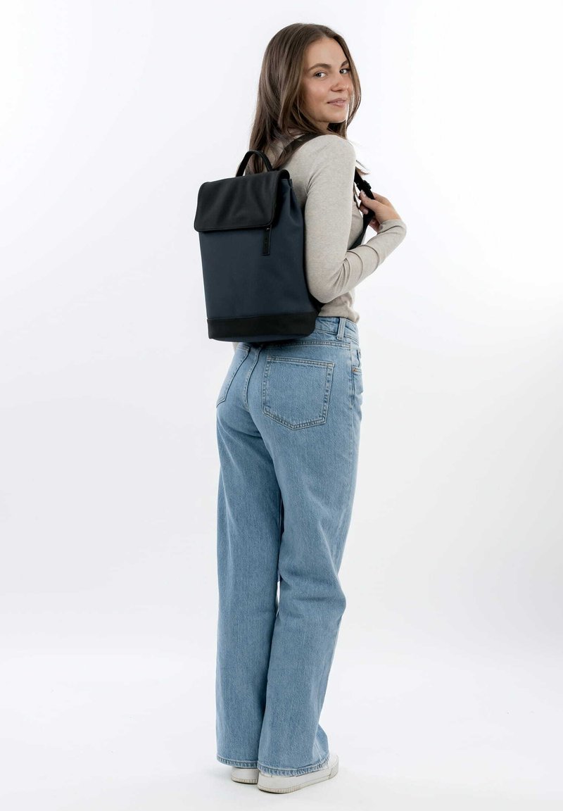 Sac à dos en tissu noir et marine avec un design à rabat, des bretelles ajustables et une finition texturée, porté sur le dos contre un fond clair.